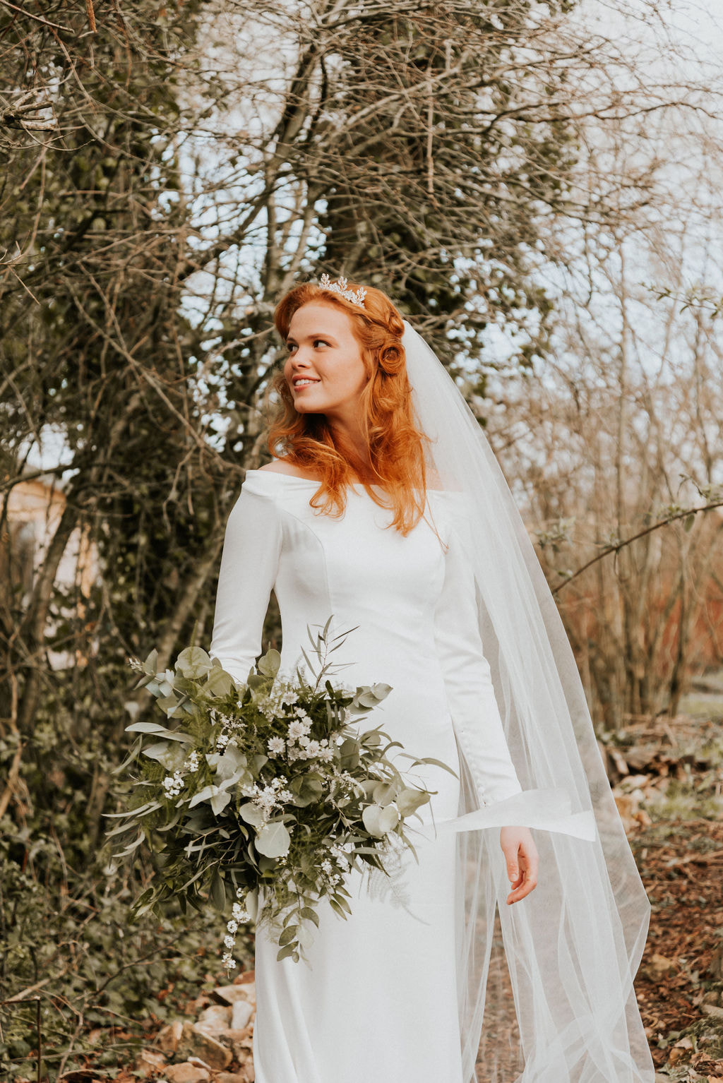 exquisite plain simple bridal long sleeve dress with long veil auburn hair and hairband with leafy bouquet in a Winter setting