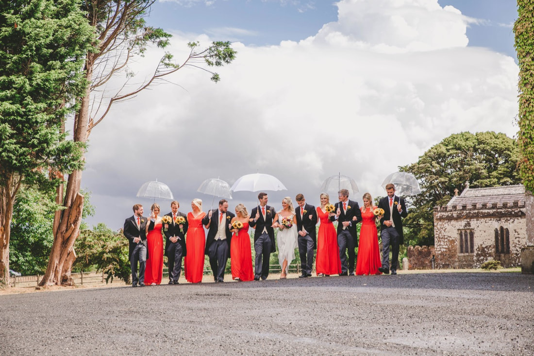group shot of wedding party, bridesmaids in bright red dresses, groomsmen in tails, umbrellas up as wedding in the rain, dramatic sky
