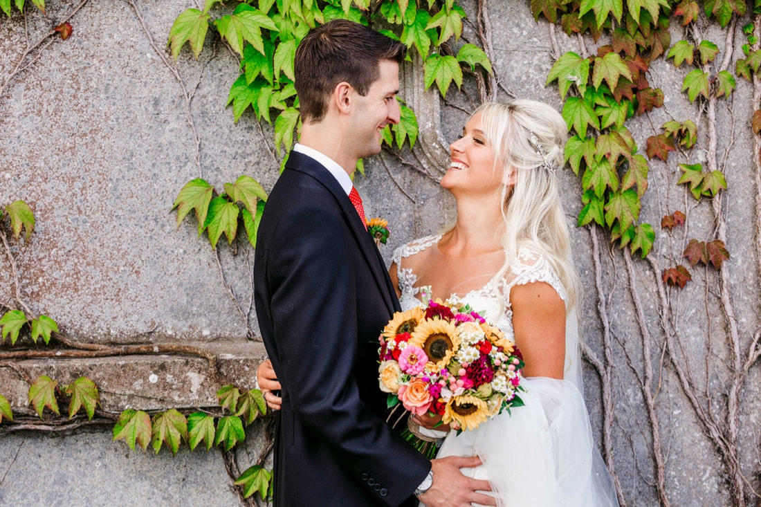 bride and groom by a wall with plants and sunflower bouquet, smiling at each other
