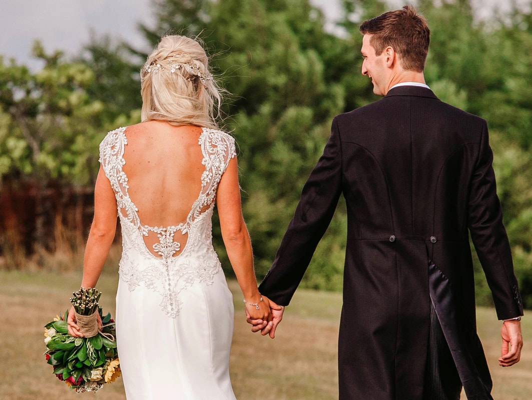 back of bride and groom walking showing wedding dress embellishment and hair accessory details, holding a bouquet, groom wearing tails