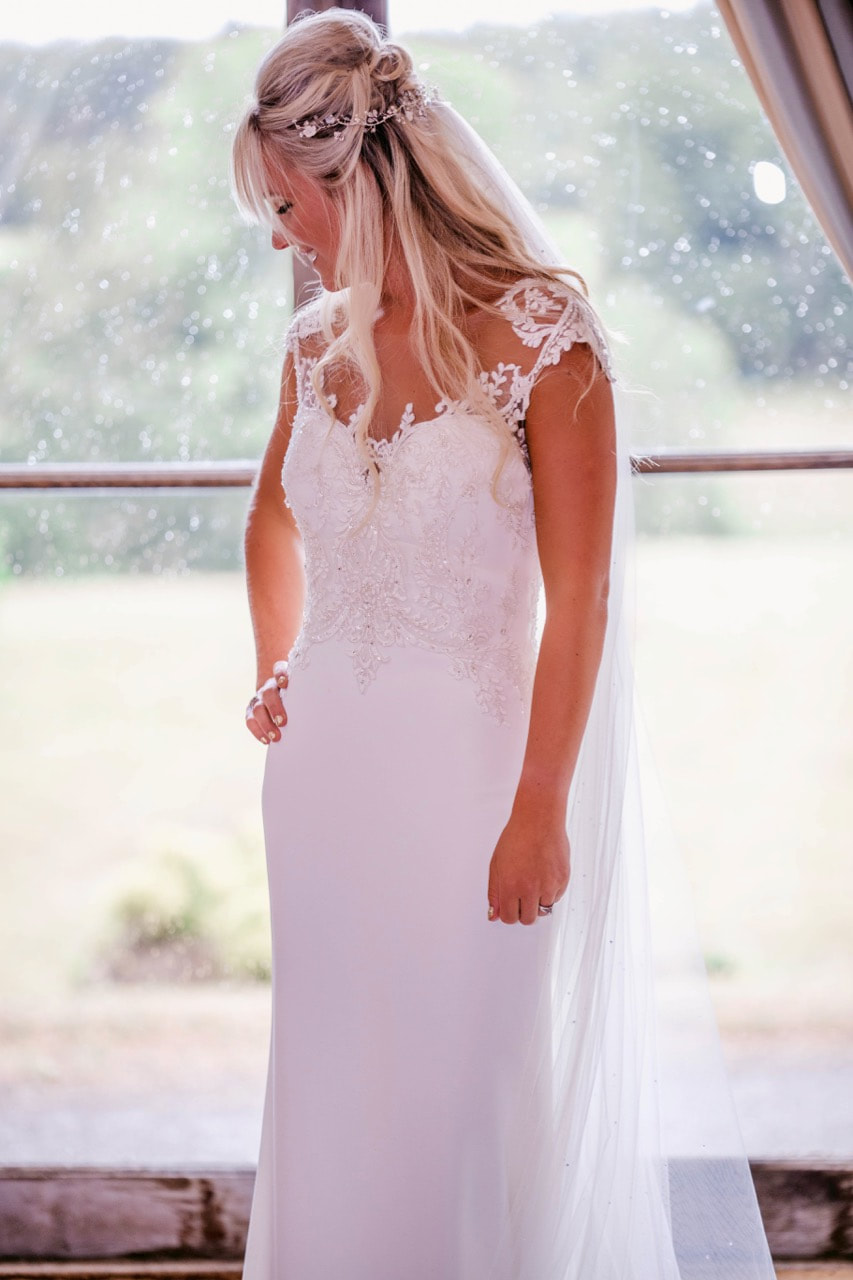 bride wearing a fitted dress with beautiful details and neckline hair half up with a delicate hairvine with mother of pearl flowers and crystals and pearls, standing by the window