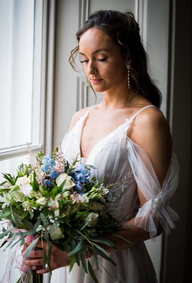 Bride standing by the window glancing at bouquet pale pink, blue and white, long drop earrings and soft floaty dress with sleeve details