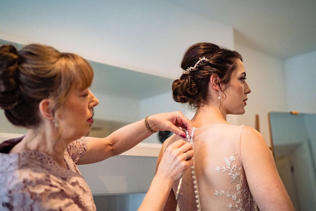 bride getting ready with her mum doing up the button on the back of her sheer back lace wedding dress, freshwater pearl earrings and hair accessory